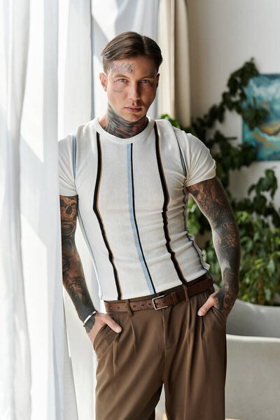 Молодой человек с татуировками уверенно стоит в помещении, демонстрируя стильный наряд у окна.