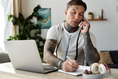 Dövmeli genç adam evde not yazarken bir telefon görüşmesi yapıyor..