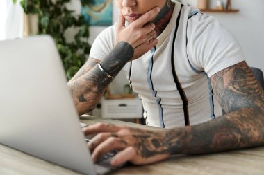 Dövmeli yakışıklı genç adam evinde bir dizüstü bilgisayarla düşünceli bir iş yapıyor..