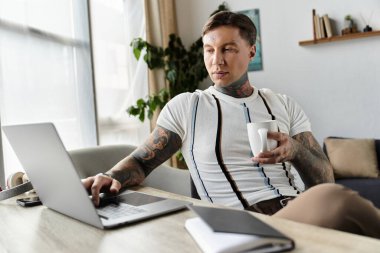 Handsome young man with tattoos focused on his laptop in a stylish and inviting room clipart