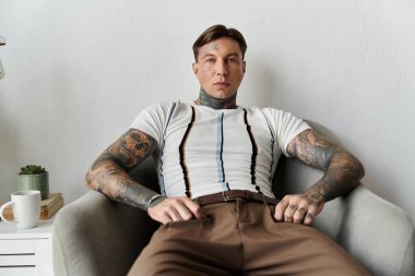 Dövmeleri olan genç bir adam rahat bir sandalyeye oturmuş, tarzını ve davranışlarını sergiliyor..