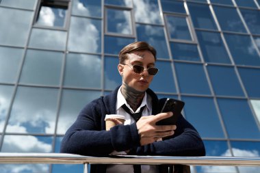 Dövmeli bir adam, cam bir yapının yakınındaki akıllı telefonuna bakarken kahveden hoşlanır..