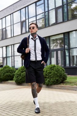 Dövmeleri olan yakışıklı genç adam çağdaş bir yapının yanında şık bir kıyafetle dolaşıyor..