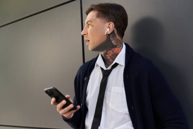 Dövmeleri olan genç bir adam telefonunda müzik dinlemekten zevk alırken bir yandan da duvara yaslanıyor..
