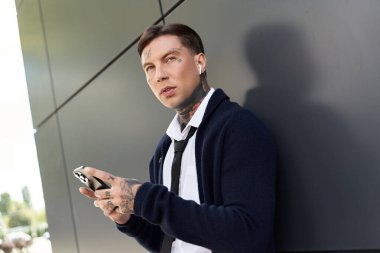 Dövmeleri olan genç bir adam dışarıda duruyor, günlük kıyafetlerini giyerken telefonuna odaklanmış..