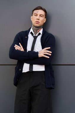 Dövmeleri olan genç adam kollarını kavuşturup çağdaş bir duvarın yanında tarzını sergiliyor..