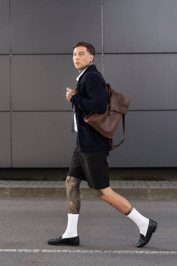 Stylish young man with tattoos strolls past modern buildings while carrying a bag. clipart