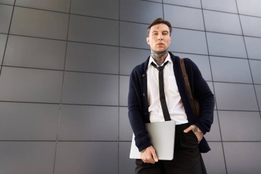 Dövmeleri olan genç bir adam elinde dizüstü bilgisayarla modern şehir ortamında poz veriyor..