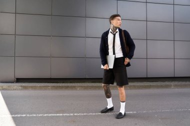 Young man with tattoos wearing stylish outfit poses confidently against urban backdrop clipart