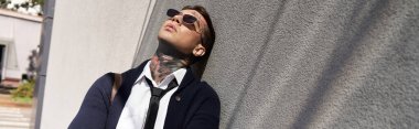 Dövmeli genç bir adam güneş ışığında gri bir duvara yaslanırken güneş gözlüğü takıyor..