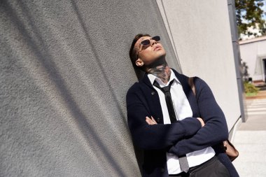 Dövmeleri olan genç bir adam duvara yaslanır, sıcak güneşin ve temiz havanın tadını çıkarır..