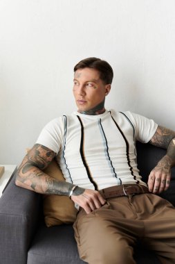 Dövmeli genç adam modern bir dairede gündelik bir koltukta oturur..