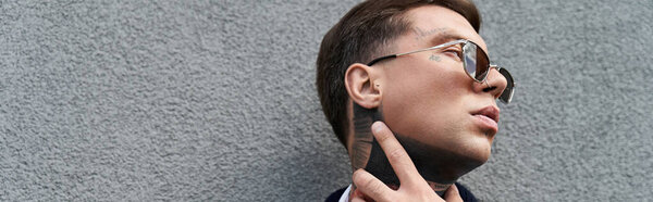 Man with tattoos and sunglasses looks thoughtfully against a gray textured wall.
