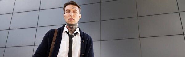 Tattooed young man in a formal outfit poses against a sleek urban backdrop.