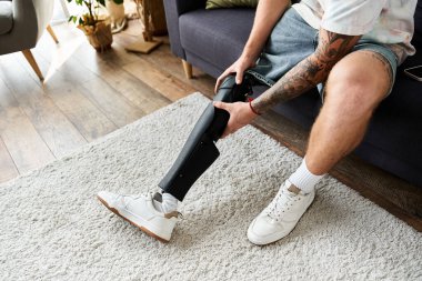 Rahat giyimli genç adam kanepeye oturur, düşünceli bir şekilde protez bacağını düzeltir..