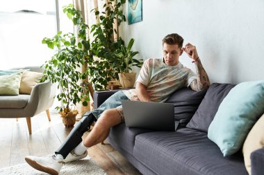 A handsome young man is seated on a modern sofa, focused on his laptop while relaxing indoors. clipart