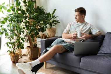 A handsome young man relaxes on a sofa, sipping coffee and using his laptop in a bright room. clipart