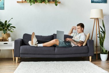 A handsome young man lounges comfortably on a cozy couch, focused on his laptop. clipart