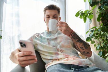 Yakışıklı genç adam rahat bir evde telefonunu karıştırırken kahvesini yudumluyor..