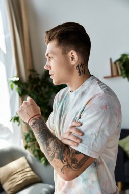 Handsome young man with tattoos gazes thoughtfully while leaning against a window in a bright room. clipart