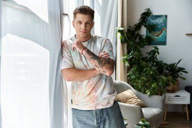 Genç bir adam, güneş ışığıyla aydınlanan bir pencerenin yanında kendine güvenerek duruyor ve tarzını gösteriyor..