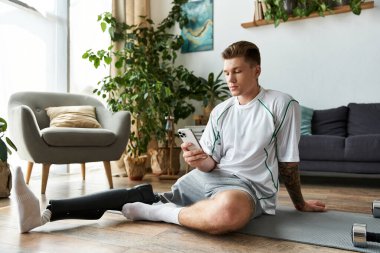 A young handsome man sits on a fitness mat in a cozy room, checking his smartphone with ease. clipart