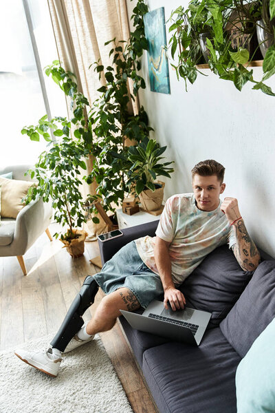 Красивый молодой человек сидит на диване с ноутбуком в яркой гостиной в окружении растений.