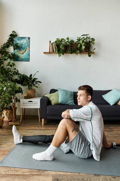 Young man with a prosthetic leg sits thoughtfully on an exercise mat in a cozy, plant filled room.