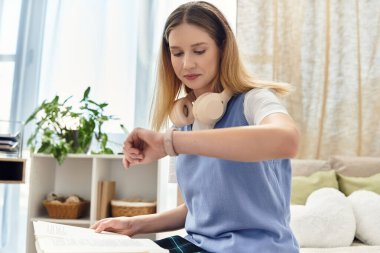 Genç bir kız evde ders çalışıyor, saatine kulaklıkla bakıyor, gençlik kültürünü kucaklıyor..