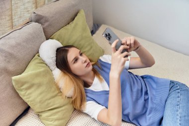 Genç bir kız odasında rahat bir şekilde uzanıyor, akıllı telefonuna odaklanmış, boş zamanlarının tadını çıkarıyor..