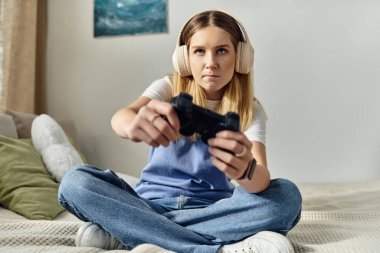 A pretty teenage girl sits cross-legged on her bed, fully focused on gaming, wearing headphones. clipart