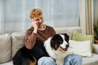 Kanepede köpeğiyle dinlenirken telefonda konuşan kızıl saçlı genç bir adam..