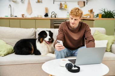 Genç adam köpeğiyle bir kanepede oturur, şık bir odada telefonuna odaklanır..
