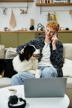 Genç kızıl saçlı bir adam köpeğini okşarken telefonla eğlenirken gülümsüyor..