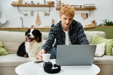 Modern bir dairede, genç kızıl saçlı bir adam, köpeği yakınlarda otururken dizüstü bilgisayarıyla rahatlar..