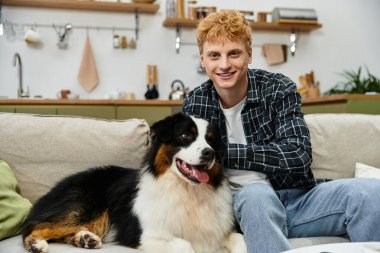 A young man with red hair relaxes on a couch, playing with his Australian Shepherd at home. clipart