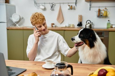 A cheerful young man with red hair talks on the phone while petting his Aussie at home. clipart