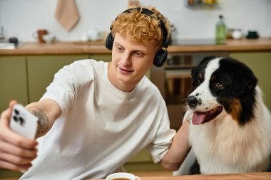 Kızıl saçlı genç bir adam, modern dairesinde Avustralyalı çobanıyla selfie çekiyor..