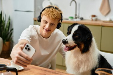 Kızıl saçlı genç bir adam, modern bir dairede şirin Avustralyalı çobanıyla selfie çekiyor..