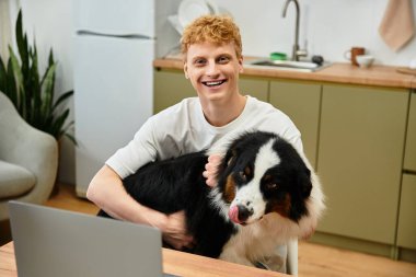 Genç adam, modern bir apartman dairesinde Avustralyalı çoban köpeğini kucaklarken gülüyor..
