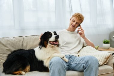 Neşeli genç kızıl saçlı adam evde telefonda konuşurken köpeğiyle rahatlıyor..