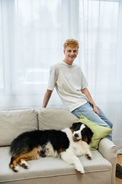 Genç kızıl saçlı bir adam rahat bir kanepede neşeli Avustralyalı çoban köpeğiyle rahatlıyor..