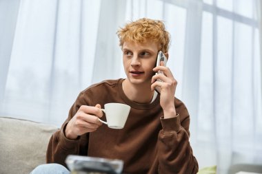 Genç, kızıl saçlı bir adam, rahat dairesinde telefonla konuşurken bir fincan içmekten zevk alıyor..