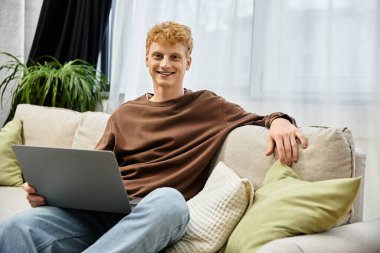 Genç, kızıl saçlı bir adam dizüstü bilgisayarıyla kanepede dinleniyor ve şık dairesinde gülümsüyor..