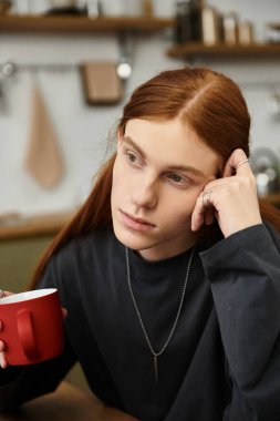 Modern bir dairede, uzun saçlı şık bir genç adam kırmızı bardaktan yudumlarken yansıyor..