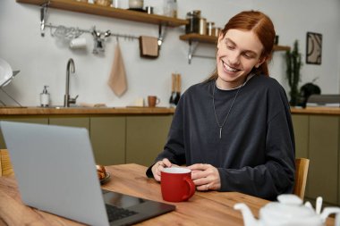 Rahat bir ortamda, uzun saçlı genç bir adam evde kahvesini yudumlarken gülümsüyor..