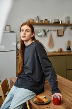 Uzun saçlı rahat bir genç adam rahat bir dairede oturuyor, yaşam tarzının tadını çıkarıyor..