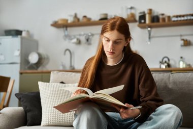 Uzun kızıl saçlı, yakışıklı genç bir adam evde kitap okuyarak rahatlıyor..