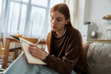 Casual young guy in a sweatshirt reflects while writing in his notebook at home. clipart