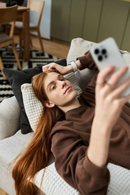 Relaxing on a cozy couch, a young redhead enjoys his time scrolling through his phone. clipart